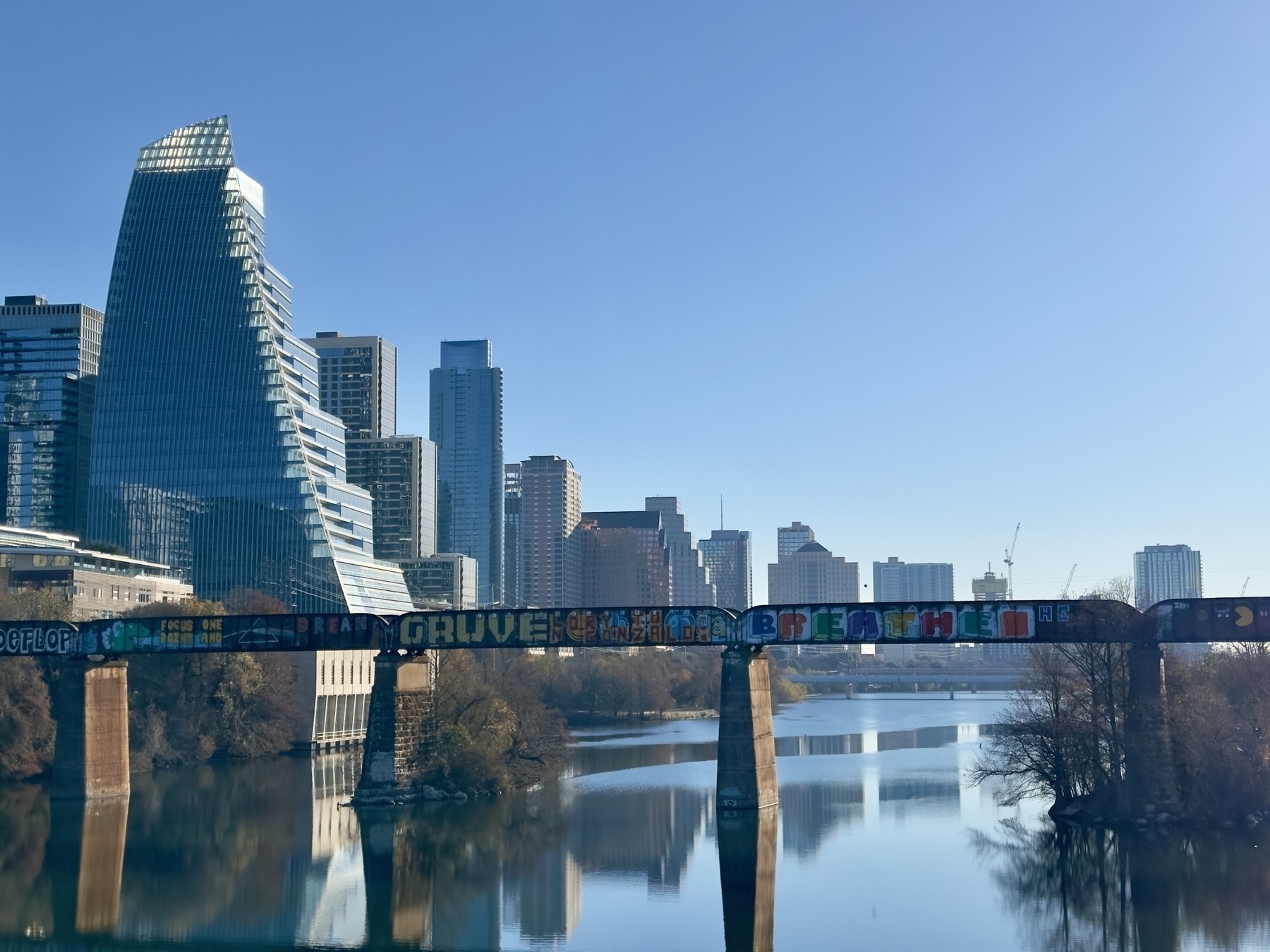 A view from the pedestrian bridge across Lady Bird lake of the Austin skyline and the graffiti covered old rail bridge. Blue skies, mirror like water, the city reflecting.